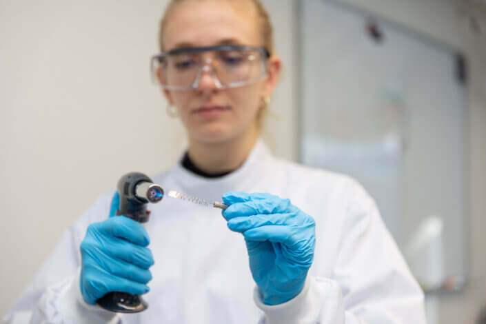 A person in a lab coat and safety goggles holds a tool in blue gloved hands and carries out an experiment in a laboratory environment.