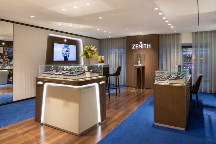 A modern watch store with showcases in which watches are displayed. The room has a wooden floor, blue carpeting and a digital screen with a watch image.