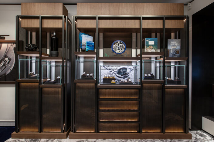 Exhibition of luxury watches in various styles in a display case made of wood and glass. The shelves above provide space for books and decorative items.