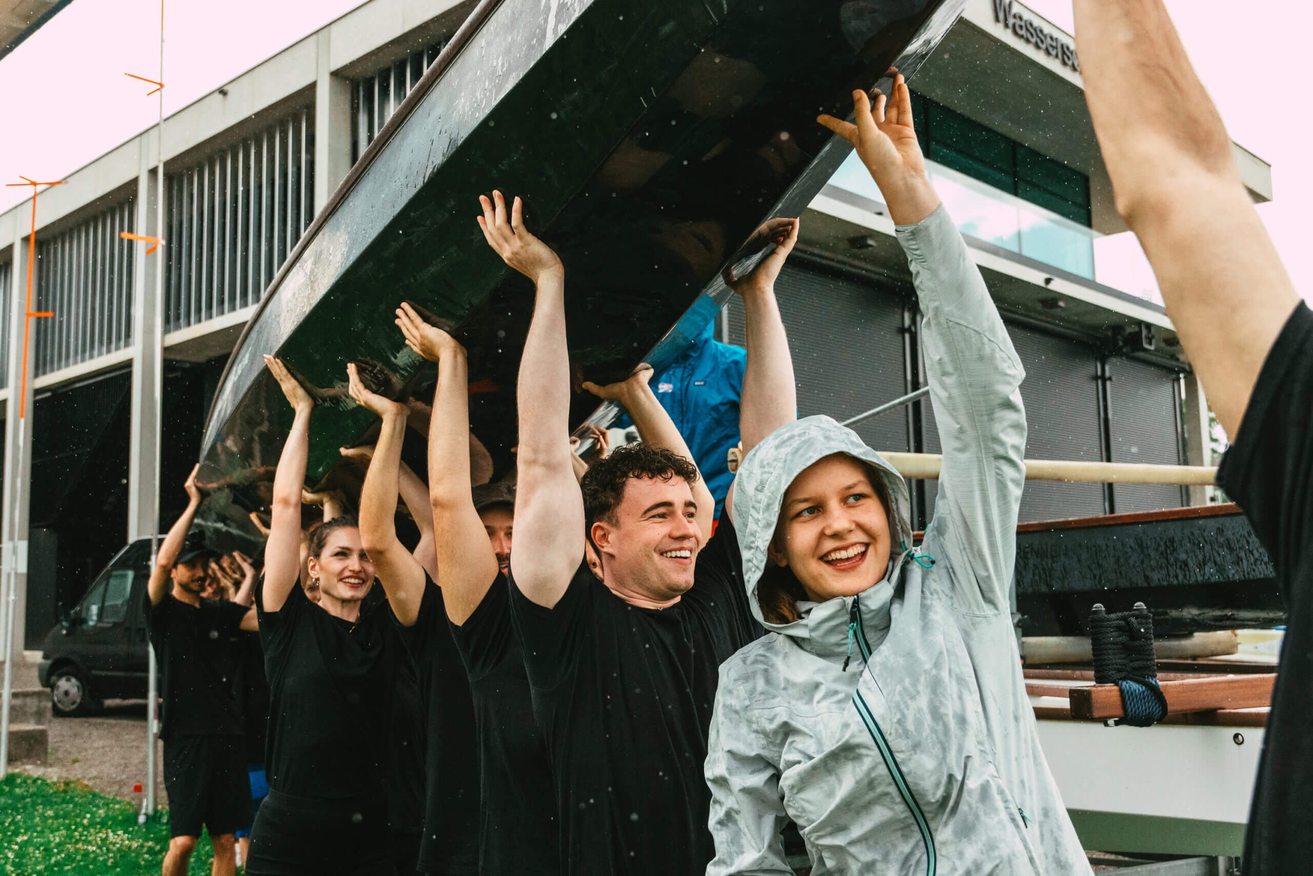 Eine Gruppe lächelnder Menschen im Regen hebt in der Nähe eines modernen Gebäudes ein Boot über den Kopf.