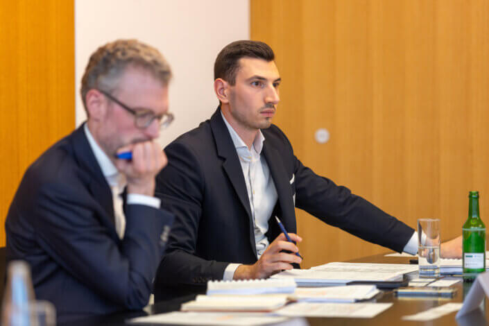 Zwei Männer in Anzügen sitzen mit Dokumenten und Wassergläsern an einem Konferenztisch. Einer schreibt mit einem Stift, der andere beugt sich nach vorne. Beide sind auf eine Diskussion oder Präsentation konzentriert.