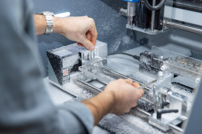 A person operates a milling machine and adjusts components while white particles swirl around. The machine is located indoors with visible gears and levers.