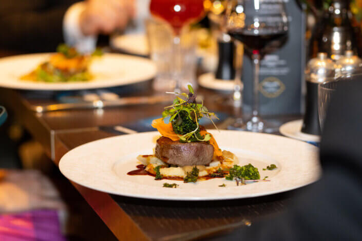 Gourmetgericht mit Fleisch, garniert mit Gemüse und Soße, serviert auf einem weißen Teller an einem Restauranttisch mit Weingläsern und anderen Gästen im Hintergrund.