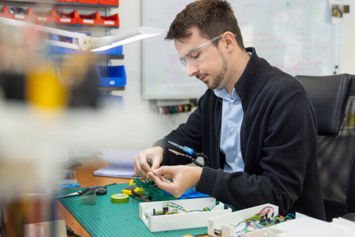 A man wearing safety goggles works on electronic devices at a desk littered with tools and cables. A whiteboard and shelves with spare parts can be seen in the background.
