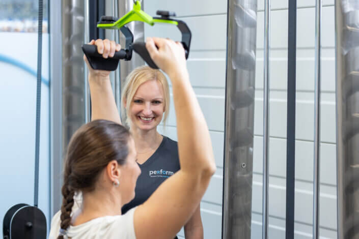 A woman in a white shirt works out on a machine while a smiling trainer instructs her in a fitness studio.