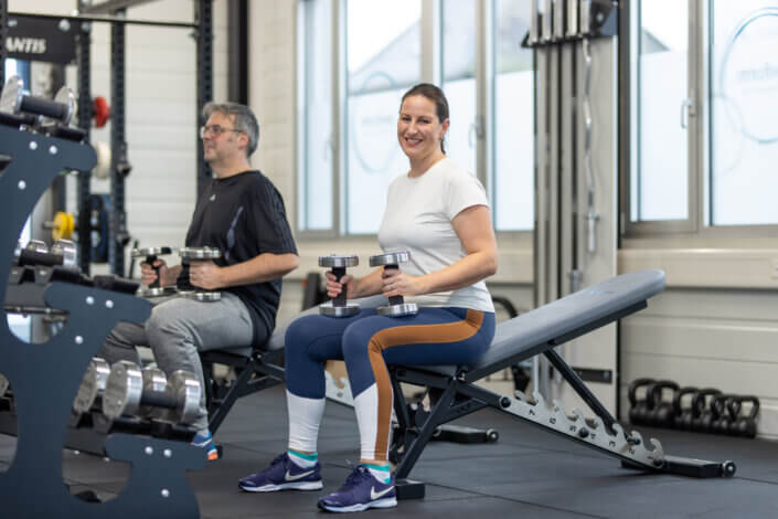 Two people are sitting on training benches in a gym, each holding dumbbells. They are standing next to each other and looking forward. Fitness equipment can be seen in the background.