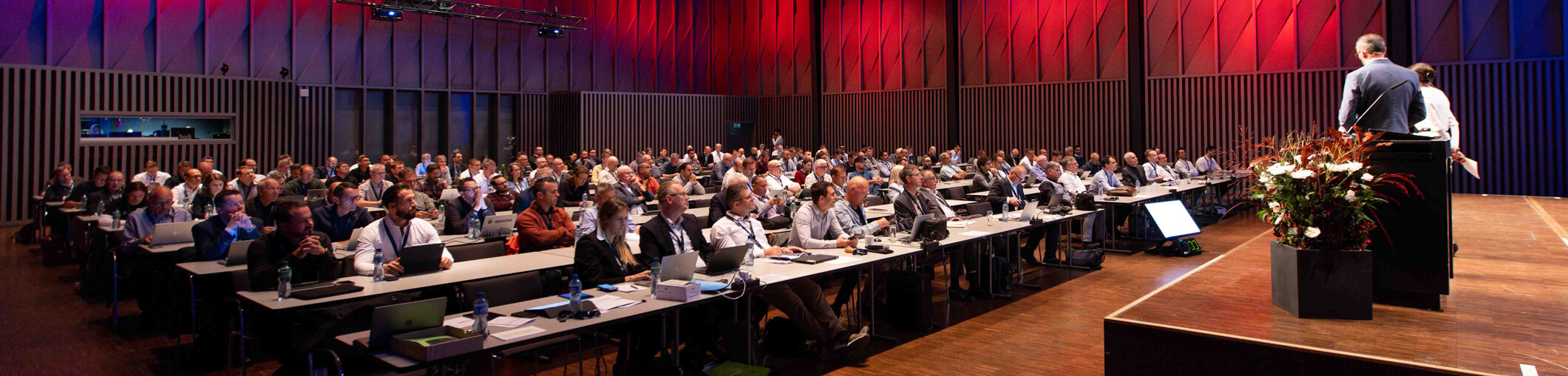 Ein Redner hält einen Vortrag vor einem großen Publikum, das an Tischen in einem Konferenzraum mit roter und blauer Beleuchtung sitzt.