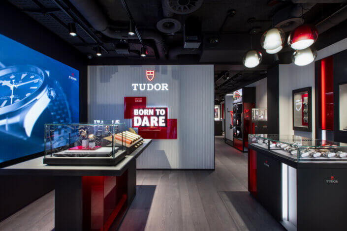 Interior view of a Tudor watch store with watches in glass cabinets and a large "Born to Dare" sign on the wall, with modern lighting and furnishings.