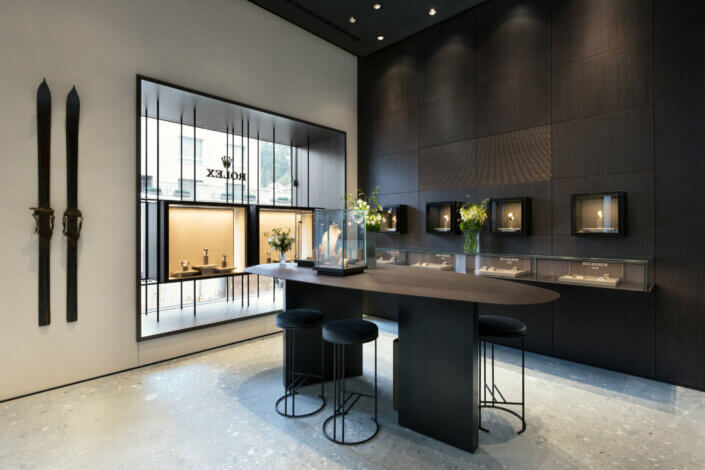 Interior of a luxury watch store with modern, black decor. On the right are showcases with watches, on the left a table with stools and skis mounted on the wall. A large window in the background.