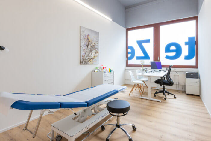 A clean, modern examination room with treatment table, stool, desk, chair, computer and large window with signage.