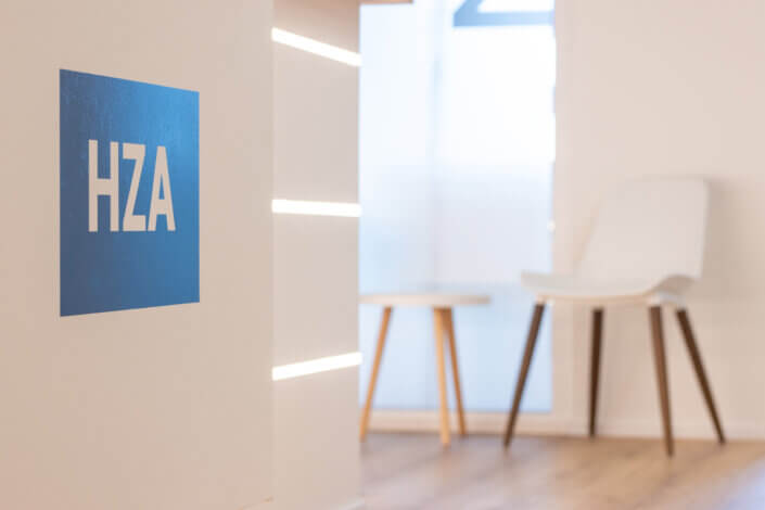 A modern interior with a blue HZA logo on a white wall, a white chair and a round wooden table on a wooden floor, all near a frosted glass door.