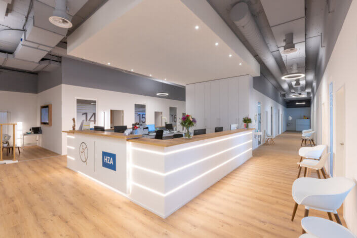 Modern office reception area with wooden floor, white walls and a central desk. Bar stools line the wall, doors with numbers can be seen in the background.