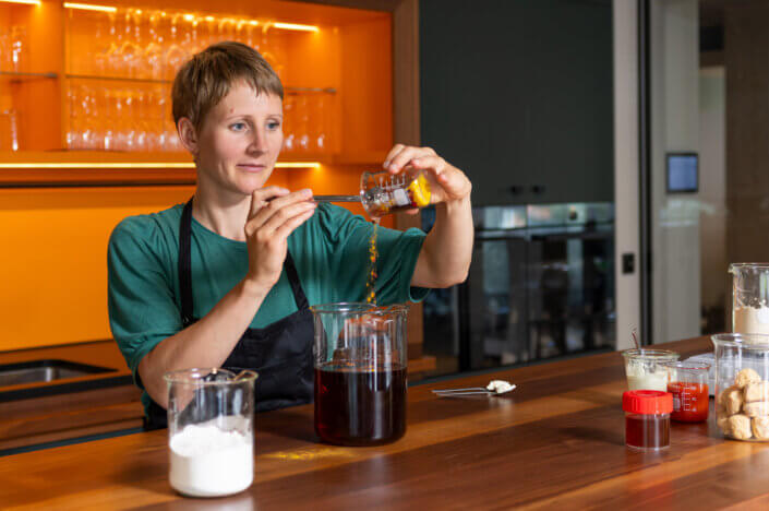 In a kitchen, a person is pouring spices into a large glass container of liquid on a wooden worktop, with jars of ingredients next to it.