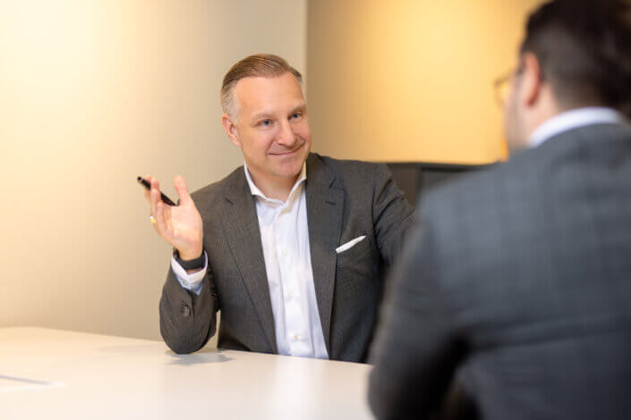 A man in a gray suit gestures with a pen while talking to another person in a meeting room.