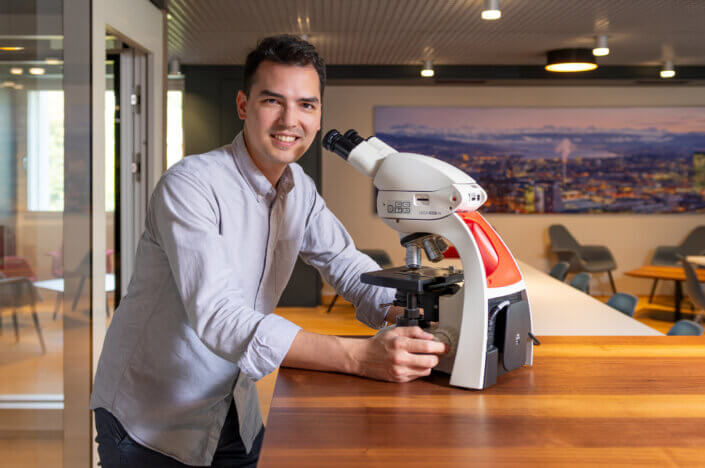 A person in a light-colored shirt stands next to a microscope on a wooden table in a modern room with a cityscape painting in the background.