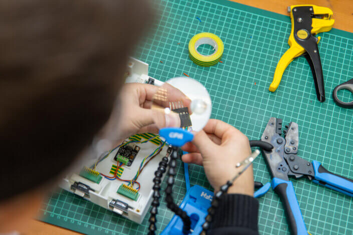 Person working on an electronic device at a table with tools and components, including cables, a magnifying glass and crimping tools, positioned on a green cutting mat.