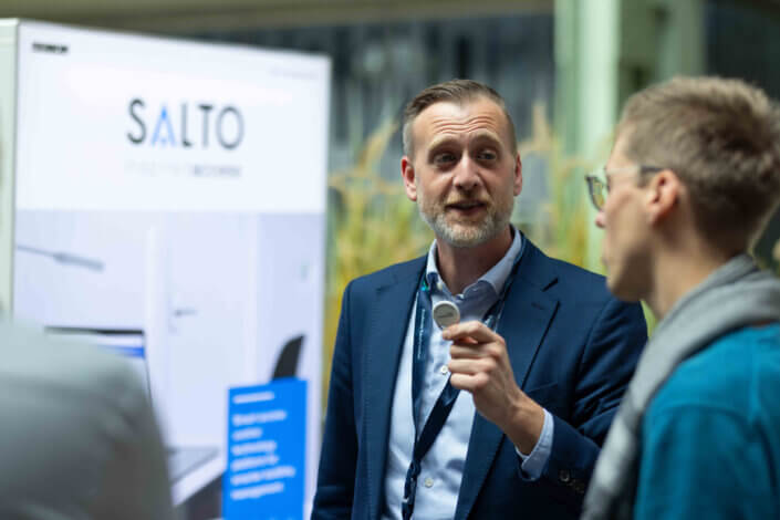 Ein Mann im Anzug spricht mit einem anderen Mann an einem Stand, im Hintergrund ist ein „SALTO“-Schild zu sehen.