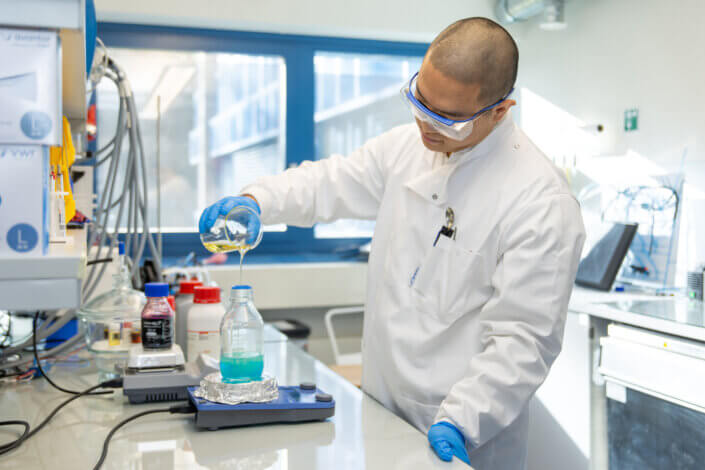 A scientist in a lab coat and safety goggles pours liquid into a flask on a hotplate in a laboratory.
