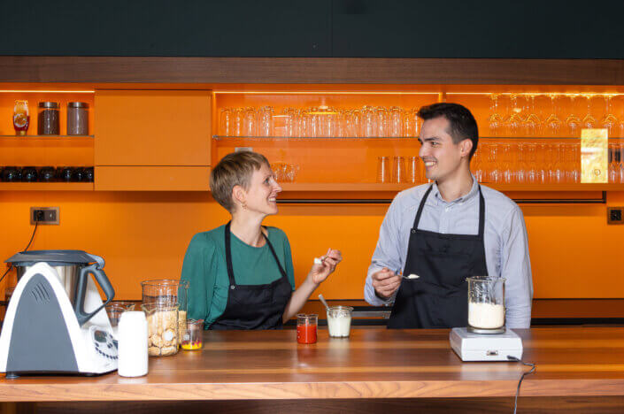 Two people in aprons stand at a kitchen counter with ingredients and utensils, smiling at each other with spoons in their hands. An orange-colored kitchen back wall and shelves with glasses can be seen in the background.