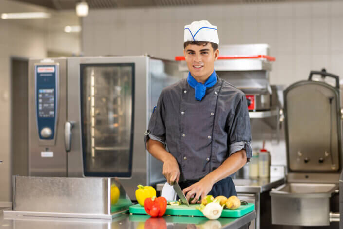 A chef in a gray uniform and white cap cuts vegetables on a green cutting board in a commercial kitchen.