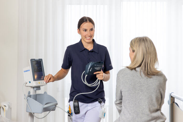 A healthcare professional with medical equipment talks to a seated woman in a clinical setting.