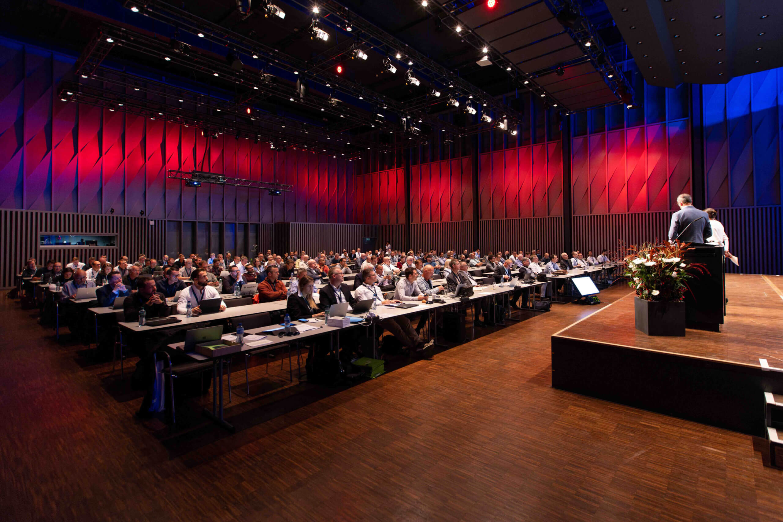 Das Publikum sitzt in einem großen Konferenzraum mit roter und blauer Beleuchtung und beobachtet einen Redner an einem Podium.