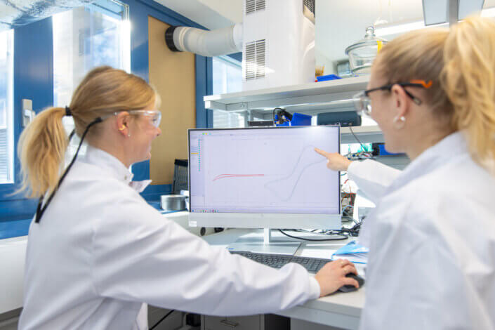 Two scientists in lab coats and protective goggles analyze a diagram on a computer screen in a laboratory environment.