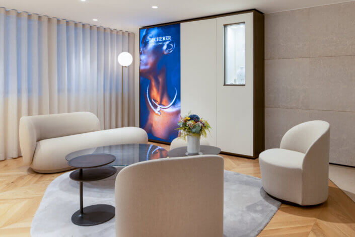 Modern lounge with white chairs and sofa, round glass table, abstract floor lamp and wall display with jewelry advertisement. Light wooden floor in herringbone pattern and transparent curtains.