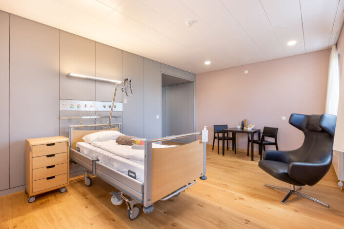 Hospital room with single bed, adjustable lamp, bedside table and black chair. There is a small table with two chairs in the corner. The room has a light-colored wooden floor and neutral walls.