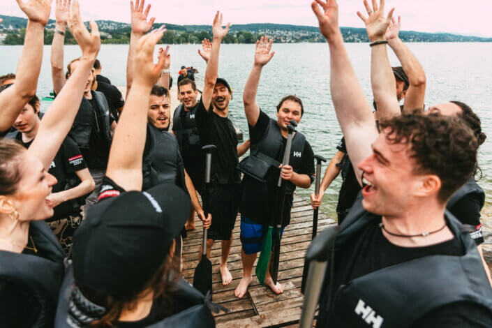 Eine Gruppe von Menschen mit Schwimmwesten und Paddeln in der Hand steht auf einem Holzsteg in der Nähe einer großen Wasserfläche, streckt die Arme in die Höhe und jubelt.