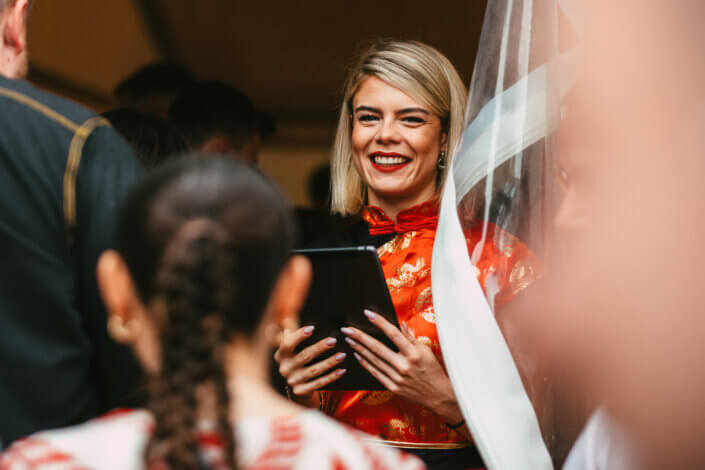 Eine Person in einem roten Kleid hält ein Tablet in der Hand und lächelt einer Versammlung zu, während sie in der Nähe eines transparenten Vorhangs steht.