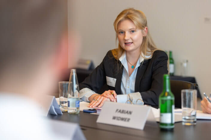 Eine Frau im Blazer sitzt an einem Konferenztisch und unterhält sich. Auf dem Tisch stehen Wasserflaschen und Namensschilder.