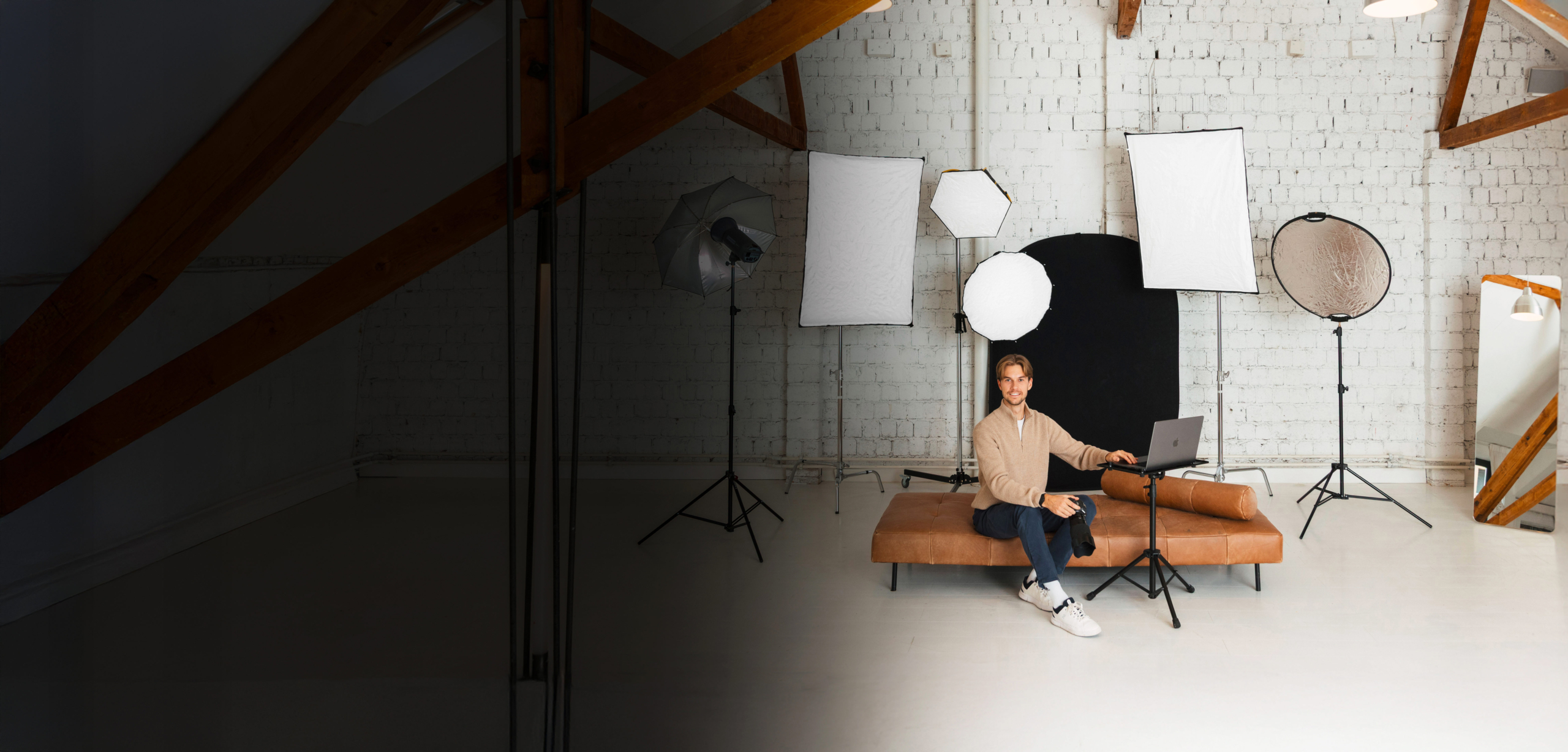 Eine Person sitzt auf einer Bank in einem Fotostudio mit Beleuchtungsausrüstung, Lichtreflektoren, einem Spiegel und einem Laptop auf einem Ständer vor einer weißen Backsteinwand.