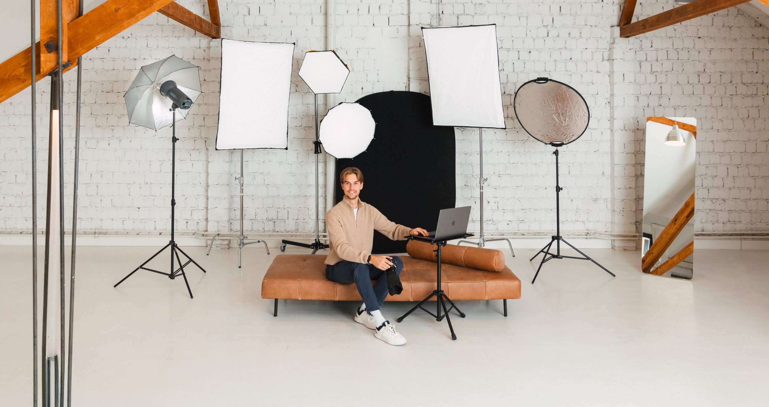 A person sits on a brown couch with a laptop in a bright studio, surrounded by several photo lamps and reflectors in front of a white brick wall. A person sits on a brown couch with a laptop in a bright studio, surrounded by several photo lamps and reflectors in front of a white brick wall.
