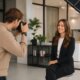 A woman in business attire is sitting there smiling while a photographer takes her picture in a modern office setting.