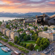 Eine Drohne fliegt bei Sonnenuntergang über das Stadtufer mit seinen Gebäuden, einem Yachthafen, Grünflächen und den Bergen in der Ferne.
