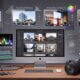 A desktop setup consisting of an iMac, a camera, headphones, memory cards, and editing software, with photo and video editing tasks displayed in German on its screens.