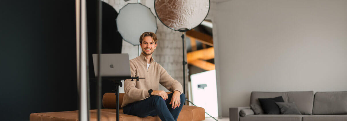Ein Mann sitzt auf einer Couch in einem modernen Studio mit Fotoausrüstung und lächelt in die Kamera, die auf einem Stativ auf einem Laptop steht.
