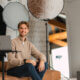 Ein Mann sitzt auf einer Couch in einem modernen Studio mit Fotoausrüstung und lächelt in die Kamera, die auf einem Stativ auf einem Laptop steht.