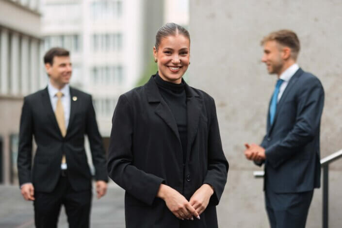 Im Vordergrund steht eine Frau in einem schwarzen Blazer und lächelt, während sich im unscharfen Hintergrund im Freien in der Nähe von Bürogebäuden zwei Männer in Anzügen unterhalten.