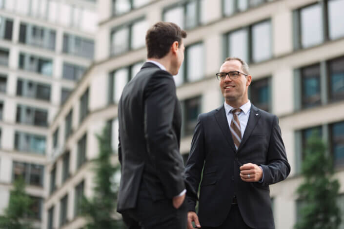 Zwei Männer in Anzügen unterhalten sich im Freien vor einem modernen Bürogebäude mit Glasfenstern.