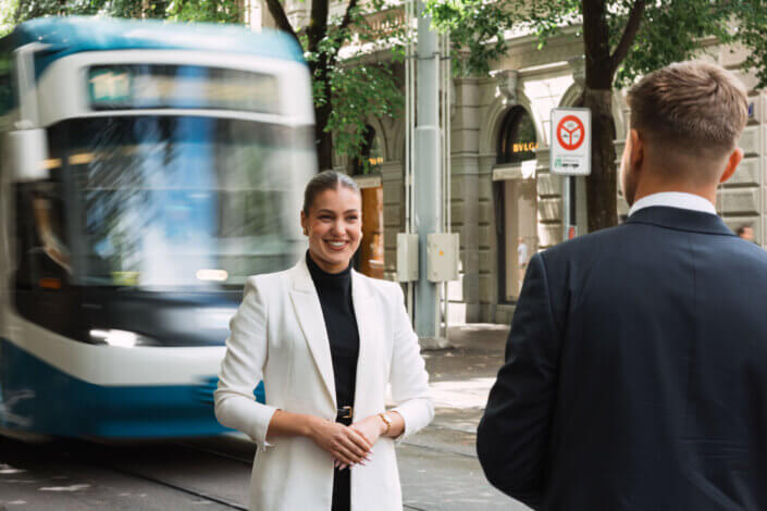Zwei Personen in Geschäftskleidung unterhalten sich auf einer Straße in der Stadt, während eine blau-weiße Straßenbahn vorbeifährt. Im Hintergrund sind Bäume und Gebäude zu sehen.