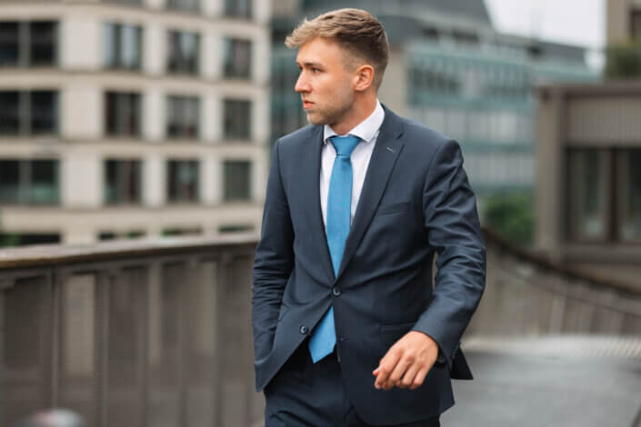 Ein Mann in blauem Anzug und Krawatte geht im Freien in der Nähe moderner Bürogebäude spazieren, blickt nach links und hat eine Hand in der Tasche.