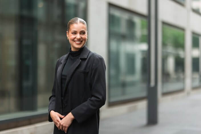 Eine Frau in einem schwarzen Blazer steht draußen in der Nähe eines modernen Gebäudes, lächelt und schaut zur Seite.